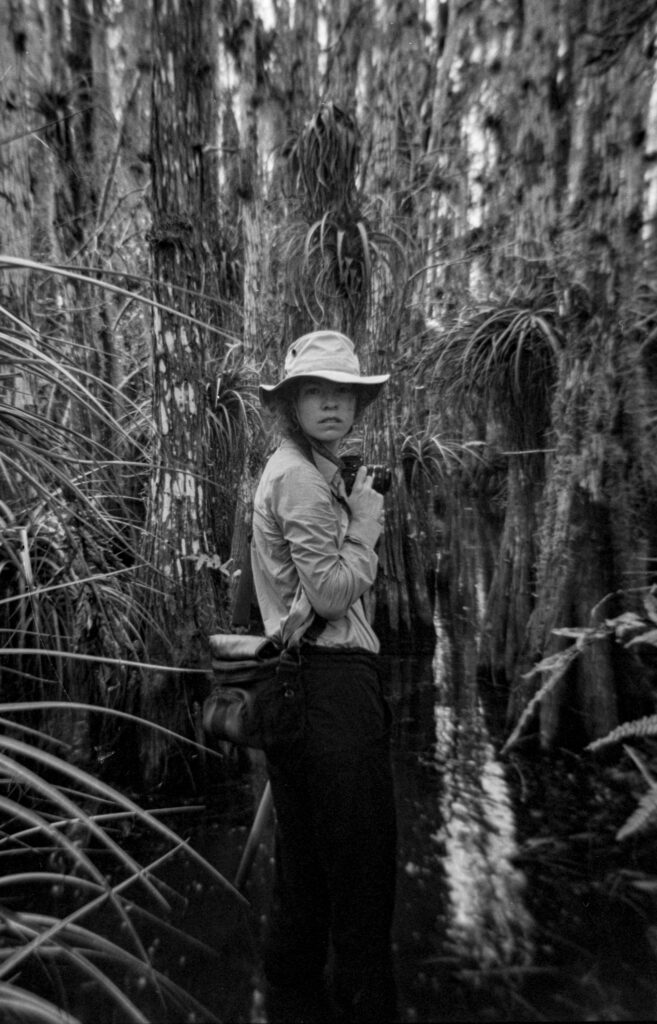 A B&W artist portrait of Rachel Gray, standing in water in a cypress tree dome with bromeliads growing throughout the trees. She holds a camera and wears a white brimmed hat.