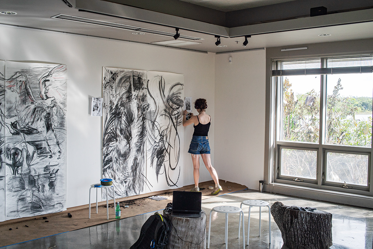 The artist, Rachel Gray, consults her reference images, as she works on her large-scale charcoal drawings of forest fires in the Everglades.