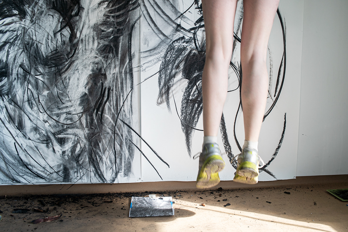 The artist, Rachel Gray, with her feet suspended mid-air, as she jumps to reach the top of her large-scale charcoal drawing. The ground below her is littered with pieces of charcoal.