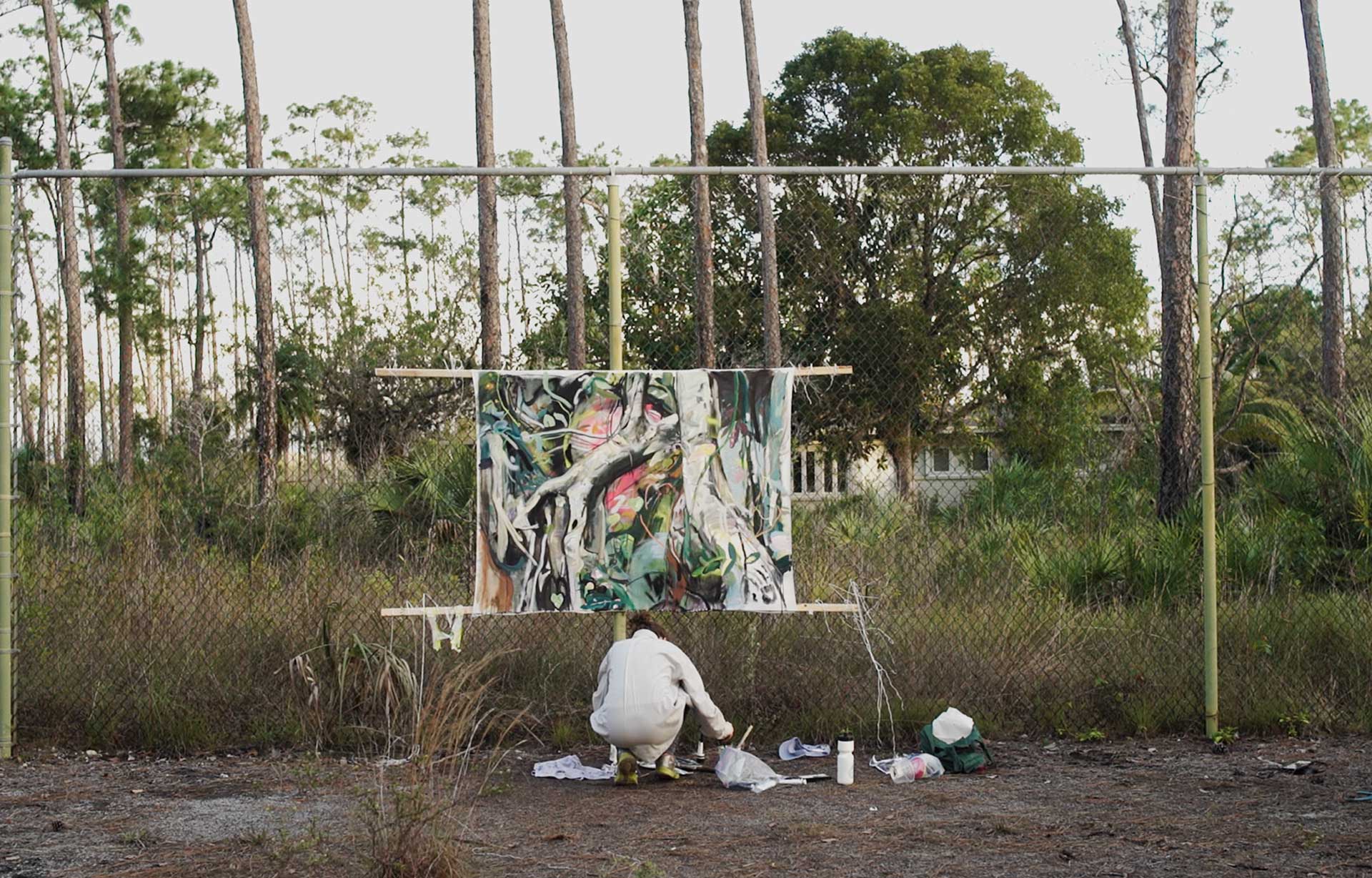 The artist, Rachel Gray, working in her outdoor studio in an abandoned basketball court. Her canvas hangs on the chain link fence. The artist is crouched before the painting as she applies paint to her brush. The painting is of a tree.