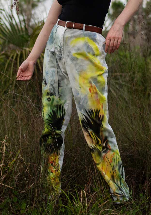 The artist, Rachel Gray, models a pair of her painted pants with paintings of fire and palm leaves.