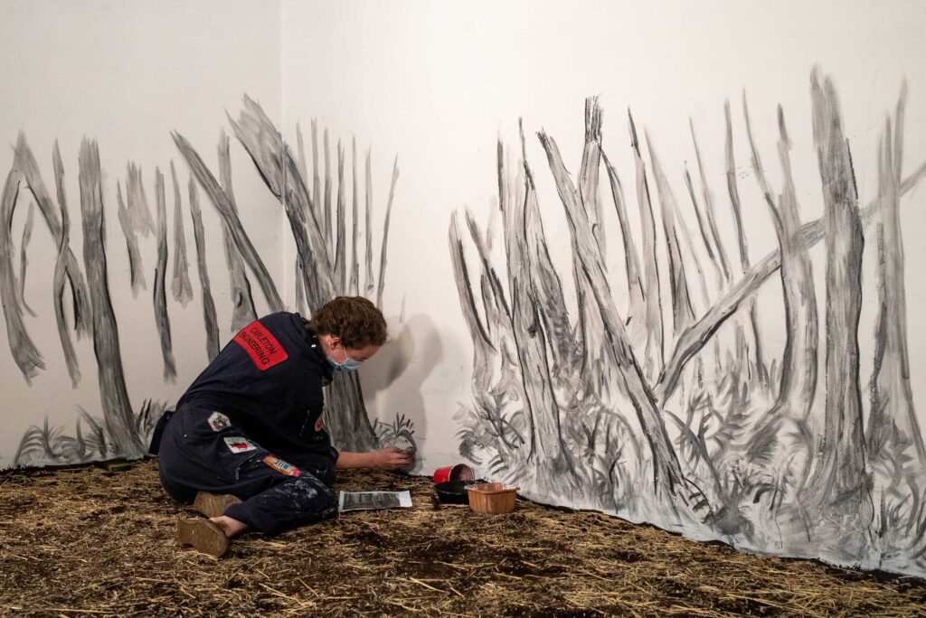 The artist, Rachel Gray, wears a blue jumpsuit and is working on a large charcoal animated. drawing of a forest for Pixie Cram's film Witch Woman.