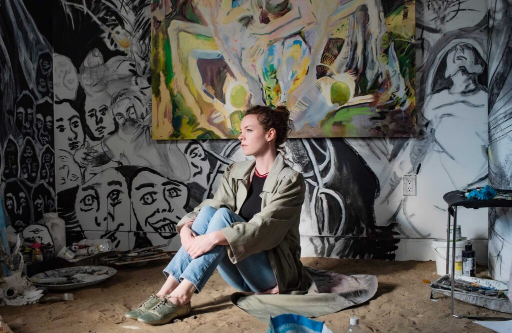 A portrait of the artist Rachel Gray in her studio. She is seated on the floor with her paintings in the background.