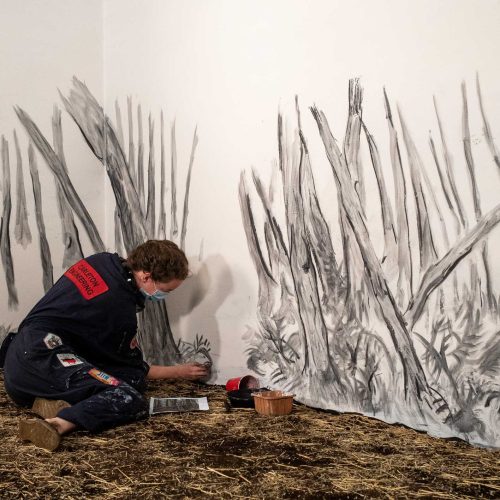 The artist, Rachel Gray, wears a blue jumpsuit and is working on a large charcoal animated. drawing of a forest for Pixie Cram's film Witch Woman.